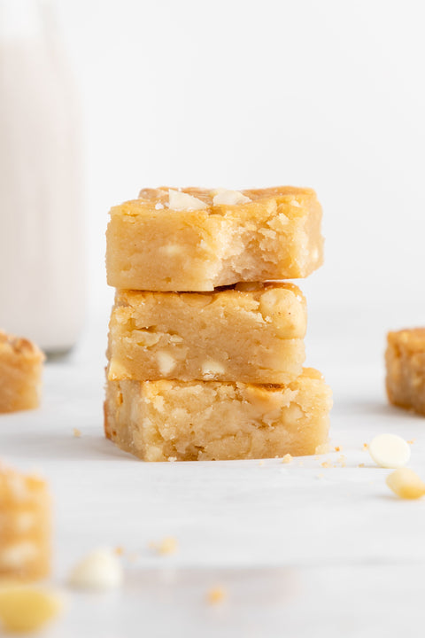 A close-up of three stacked white chocolate macadamia nut blondies on a white surface. The blondies are golden, dense, and moist with visible chunks of macadamia nuts and white chocolate chips. A bite is taken out of the top piece.