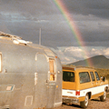 A vintage silver Airstream trailer hitched to a retro yellow and white SUV parked in a scenic desert landscape. A vibrant rainbow arcs across a moody sky, with mountains in the distance under dramatic clouds.