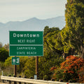 photo of a street sign on a freeway with mountains and trees in the background that says downtown right, carpinteria state beach. 